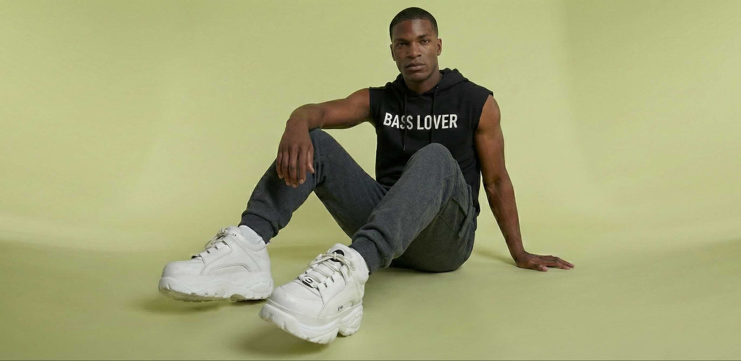 Man sitting on a green floor wearing a black 'BASS LOVER' hoodie and white sneakers.